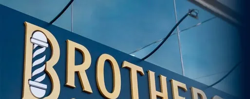 Letreiro BROTHERS Barbearia em letras douradas sobre fundo azul, com o poste de barbeiro no B